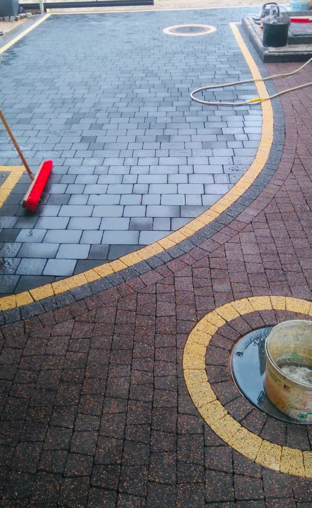 Świeżo ułożona, wilgotna kostka brukowa w dwóch odcieniach szarości i brązu, oddzielona żółtym pasem. Widoczny czerwony mop i wiadro po impregnacie. W tle widoczny sprzęt do czyszczenia.