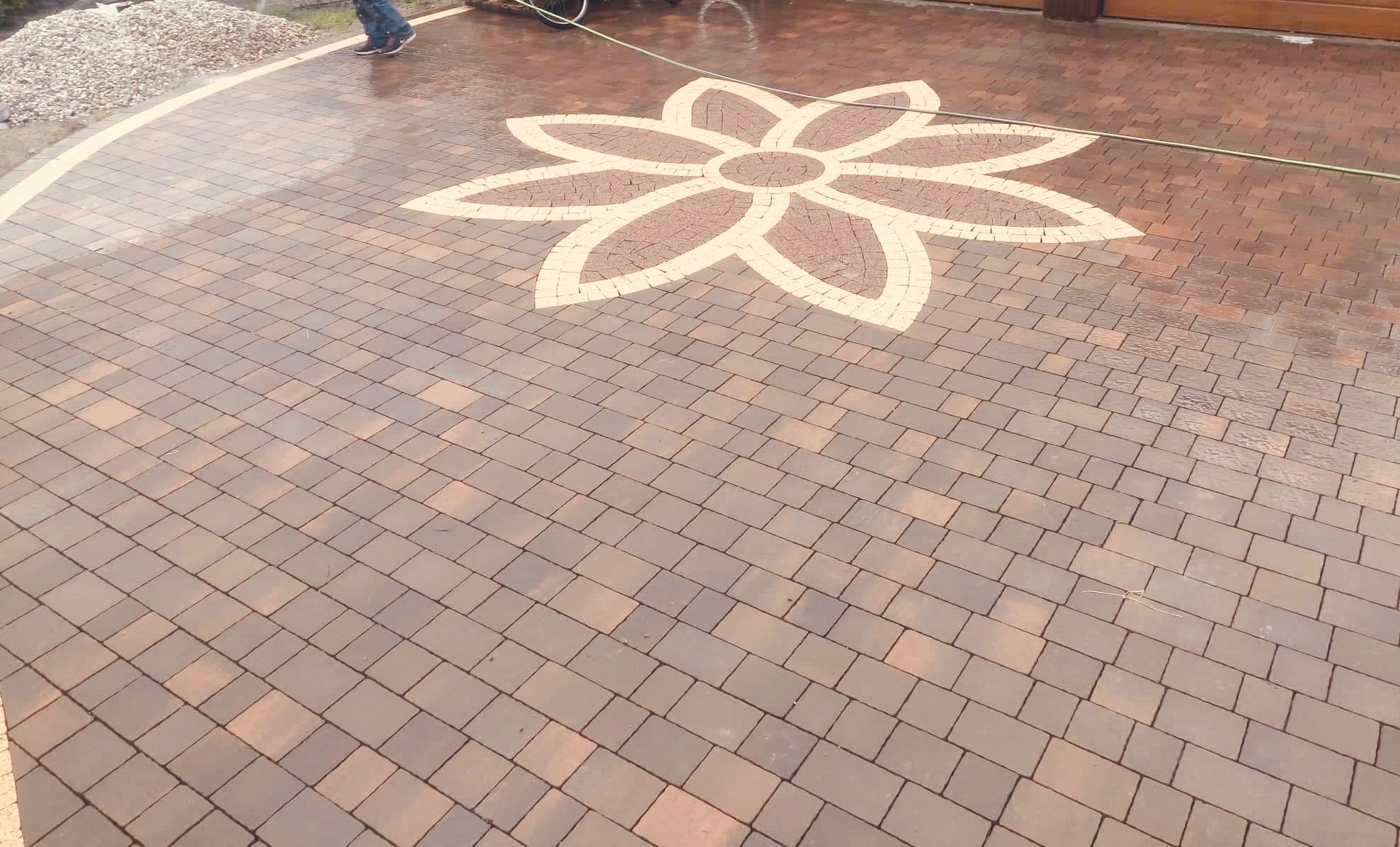 Umyta kostka brukowa w odcieniach brązu z dekoracyjnym wzorem kwiatowym w jaśniejszym odcieniu, ułożona na podjeździe przed garażem.