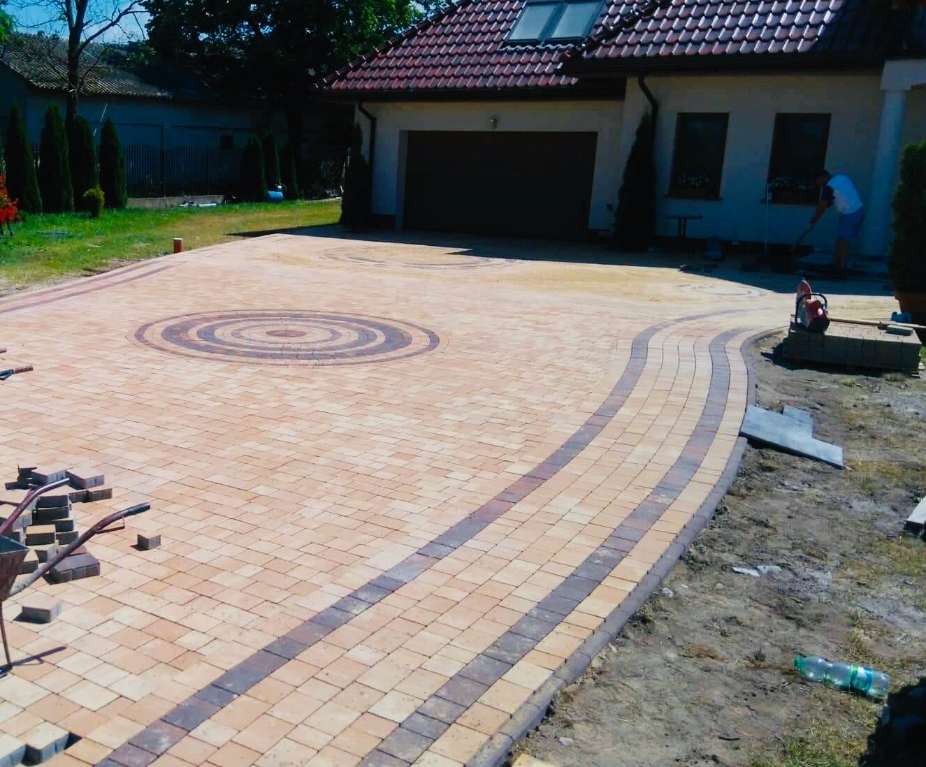 Ułożona beżowo-brązowa kostka brukowa na podjeździe z dekoracyjnym okręgiem. W tle dom z czerwoną dachówką i osoba pracująca w ogrodzie. Widoczna zagęszczarka i taczka z narzędziami.