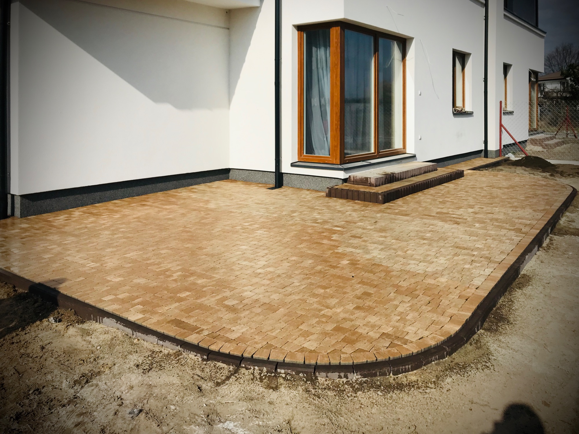 Ułożona kostka brukowa w kolorze piaskowym przed nowoczesnym domem z białymi ścianami i brązowymi ramami okiennymi, widoczne schody z kostki brukowej prowadzące do drzwi tarasowych.