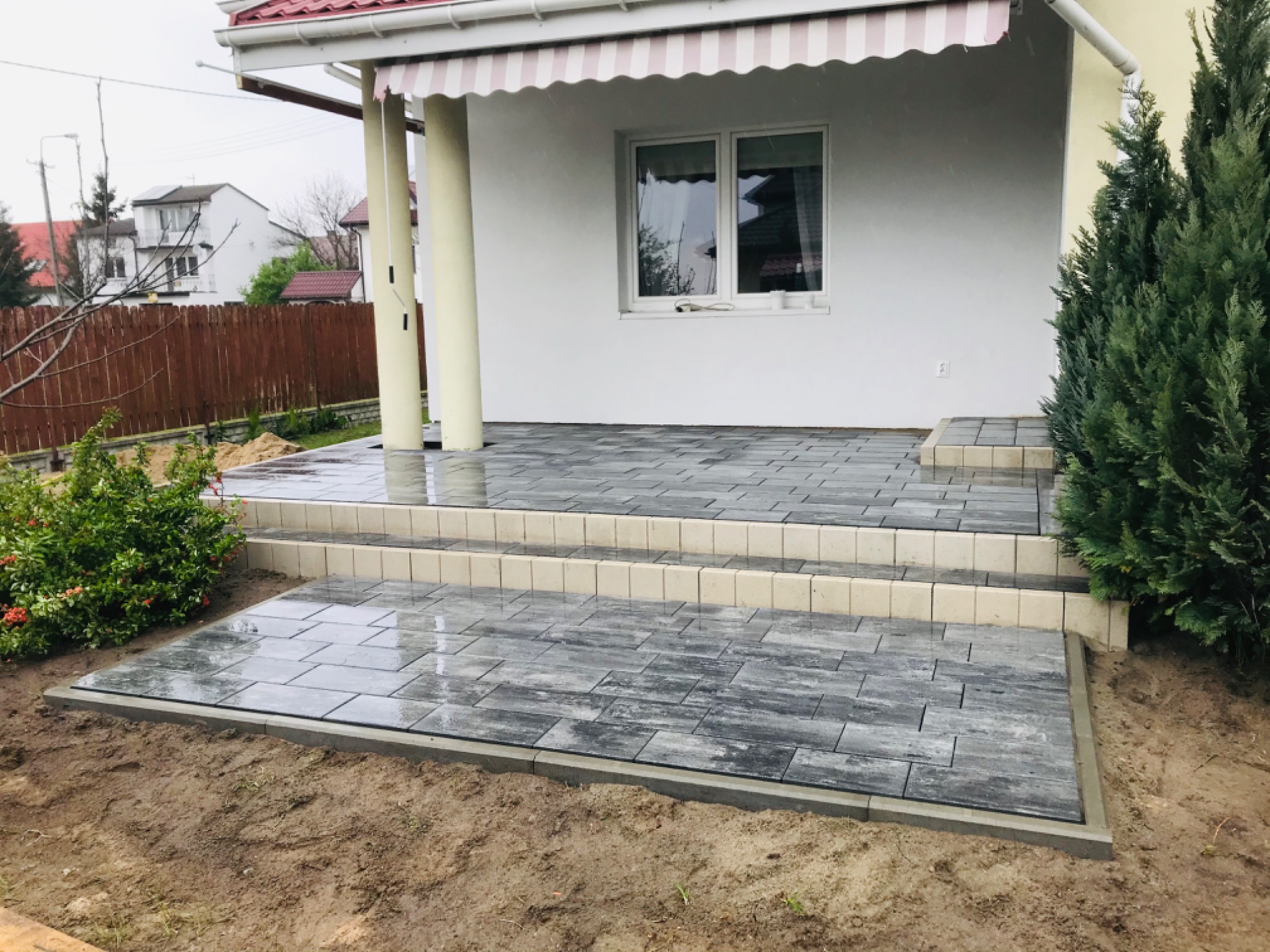 Nowo ułożony taras z szarej kostki brukowej imitującej drewno, z dwoma stopniami, prowadzący do domu z białymi ścianami i markizą, w pochmurny dzień.
