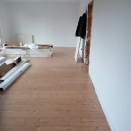 Jasne panele podłogowe z widocznym wzorem słojów, ułożone w dużym, pustym pokoju z białymi ścianami. W tle stos paneli i grzejnik. Ościeżnica drzwi bez drzwi. Ubranie wisi na drzwiach.