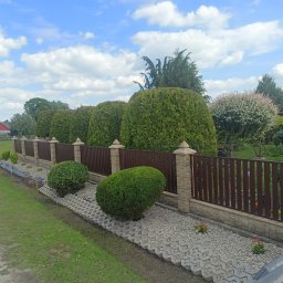 STALOVE PROFESJONALNY MONTAŻ OGRODZEŃ ARKADIUSZ DUDEK - Nowoczesne ogrodzenie panelowe z brązowymi przęsłami i kamienną podmurówką. Wzdłuż ogrodzenia posadzone kuliste tuje i ozdobne krzewy. Widok na błękitne niebo z chmurami.