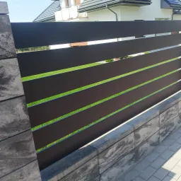 Nowoczesne ogrodzenie z poziomych, grafitowych paneli na betonowej podmurówce. W tle widoczny dom z białymi ścianami i szarym dachem.