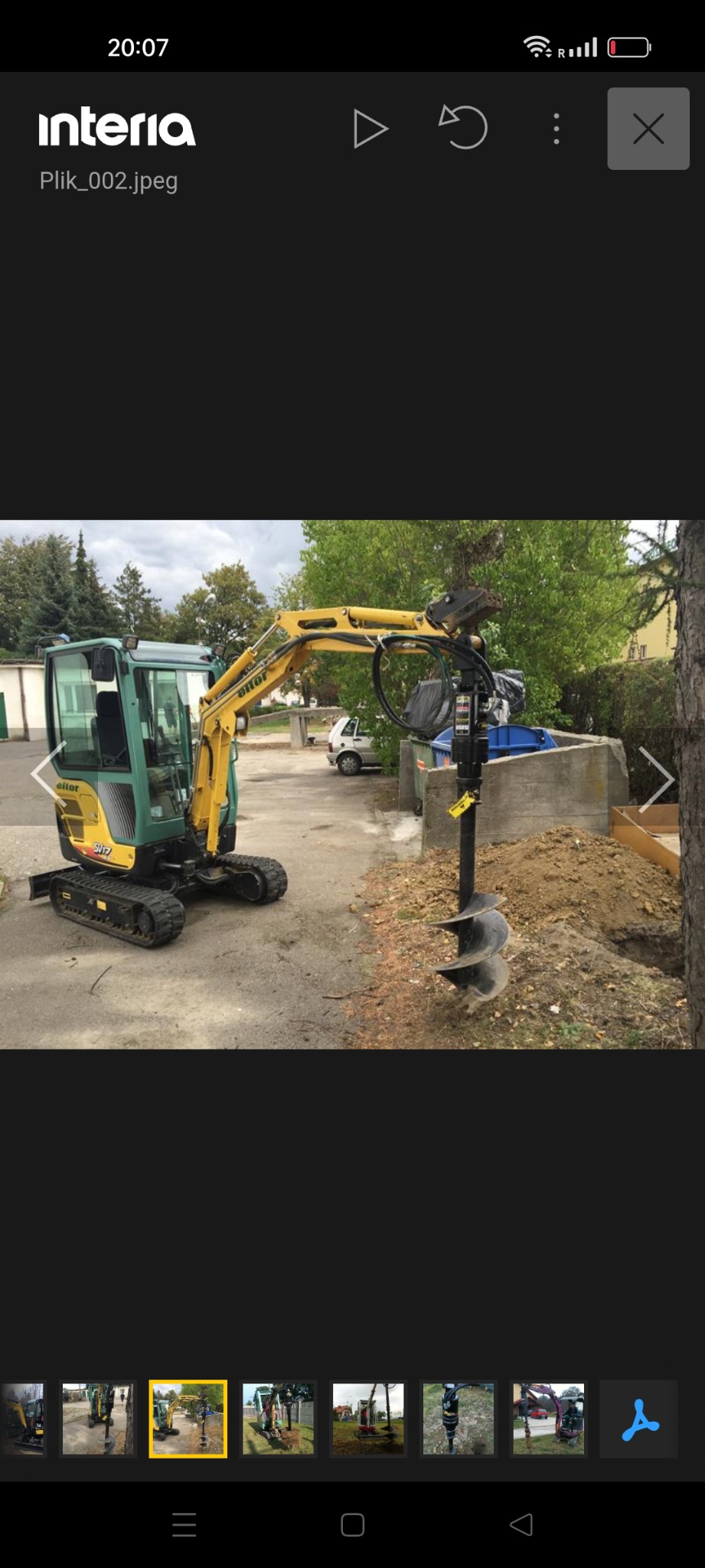 Żółto-zielona mini koparka z wiertnicą w trakcie wiercenia otworu w ziemi. W tle widoczne drzewa i budynek. Etap przygotowania pod fundament ogrodzenia.