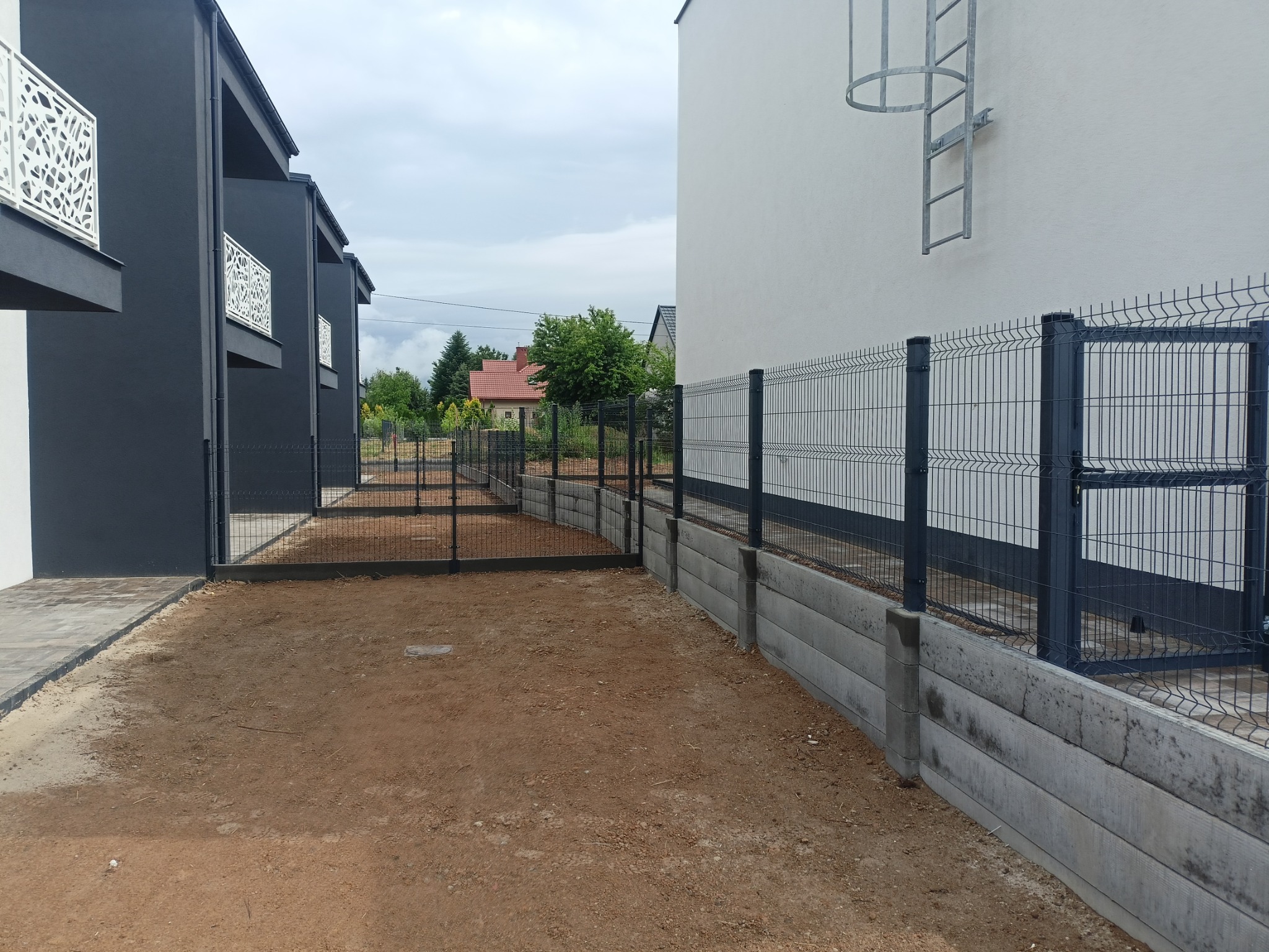 Nowoczesne ogrodzenie panelowe z podmurówką w Połańcu, widok z zewnątrz posesji. Ciemna kolorystyka paneli i słupków, betonowa podmurówka, w tle budynki mieszkalne.
