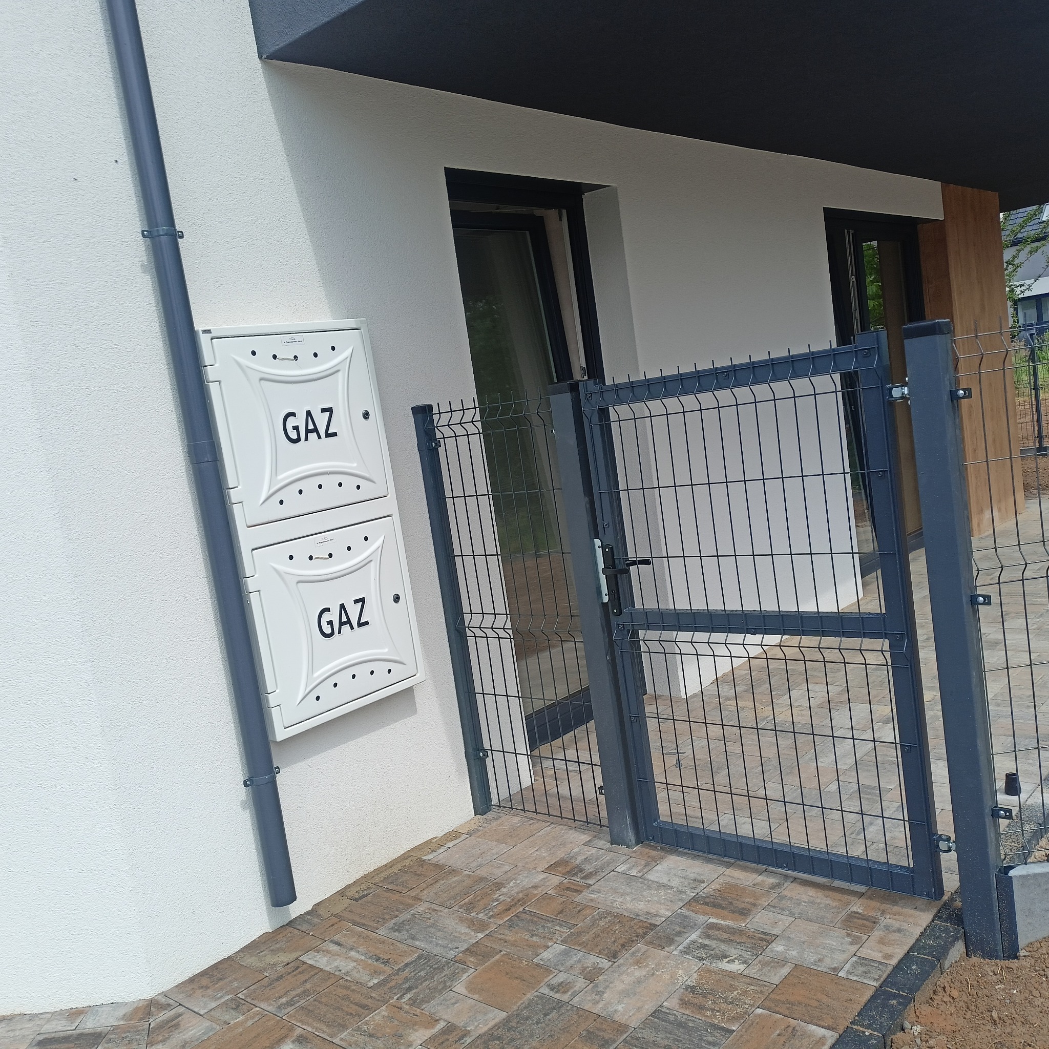 Nowoczesna furtka panelowa w kolorze grafitowym, zamontowana przy budynku z białą elewacją i skrzynką gazową. Widoczna kostka brukowa i fragment ogrodzenia panelowego.