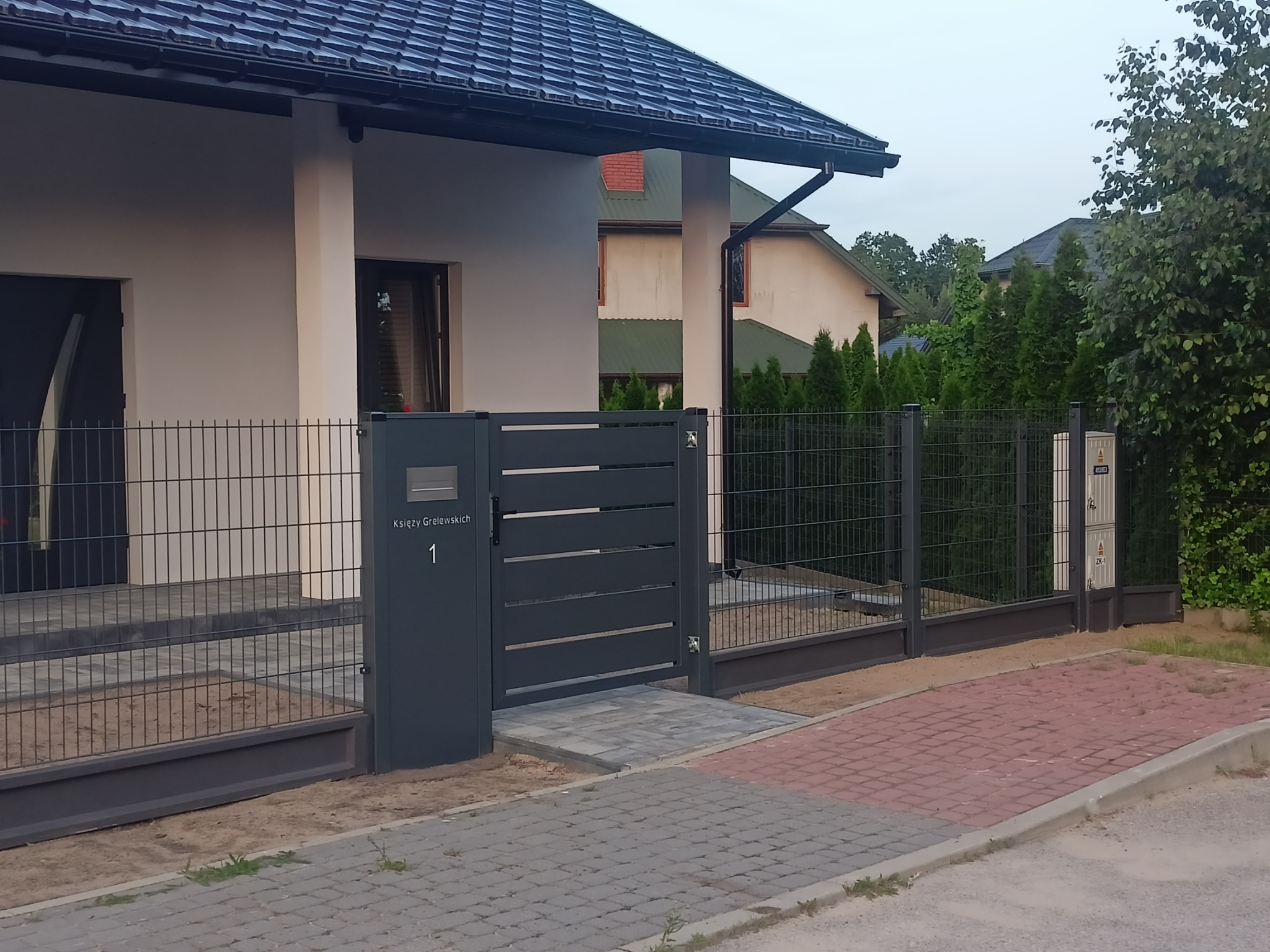 Nowoczesne ogrodzenie panelowe w kolorze grafitowym z furtką i skrzynką na listy, posesja z kostki brukowej, dom jednorodzinny w tle.