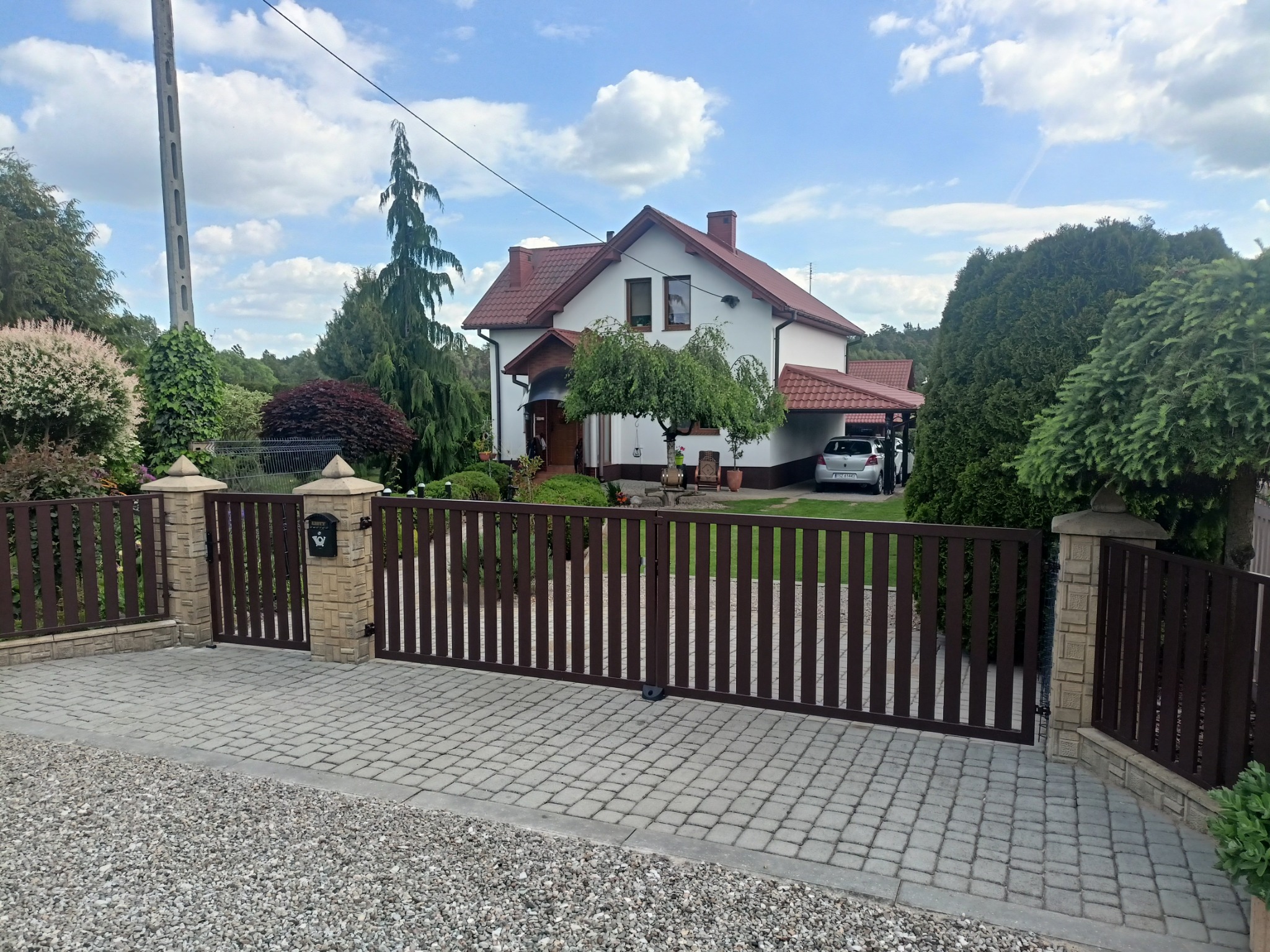 Nowoczesna, brązowa brama wjazdowa z pionowymi szczebelkami, osadzona między murowanymi słupkami, na tle domu z czerwonym dachem i zadbanego ogrodu.