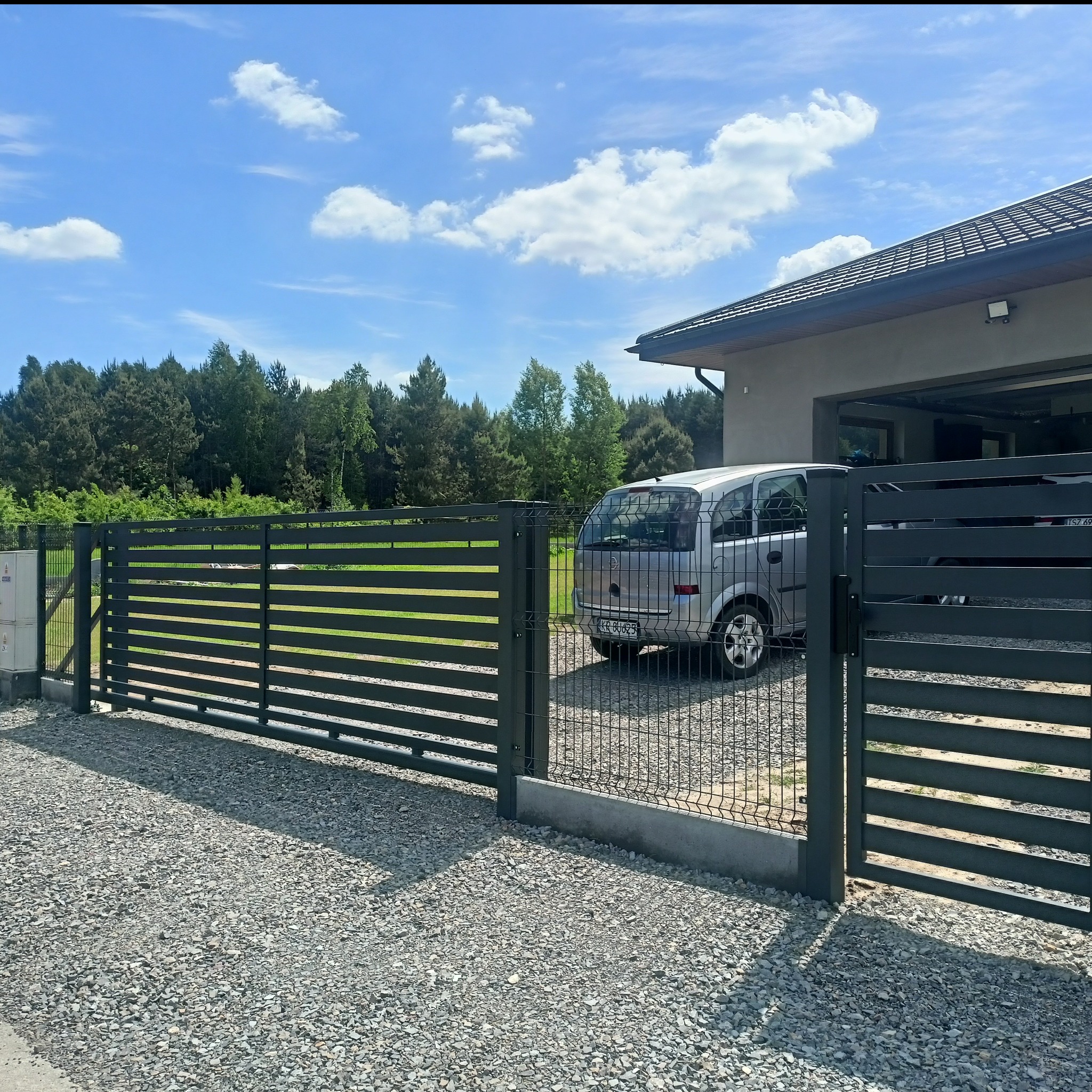 Nowoczesna, przesuwna brama wjazdowa z poziomych profili, w kolorze grafitowym, z widoczną furtką obok. W tle dom jednorodzinny i samochód osobowy.