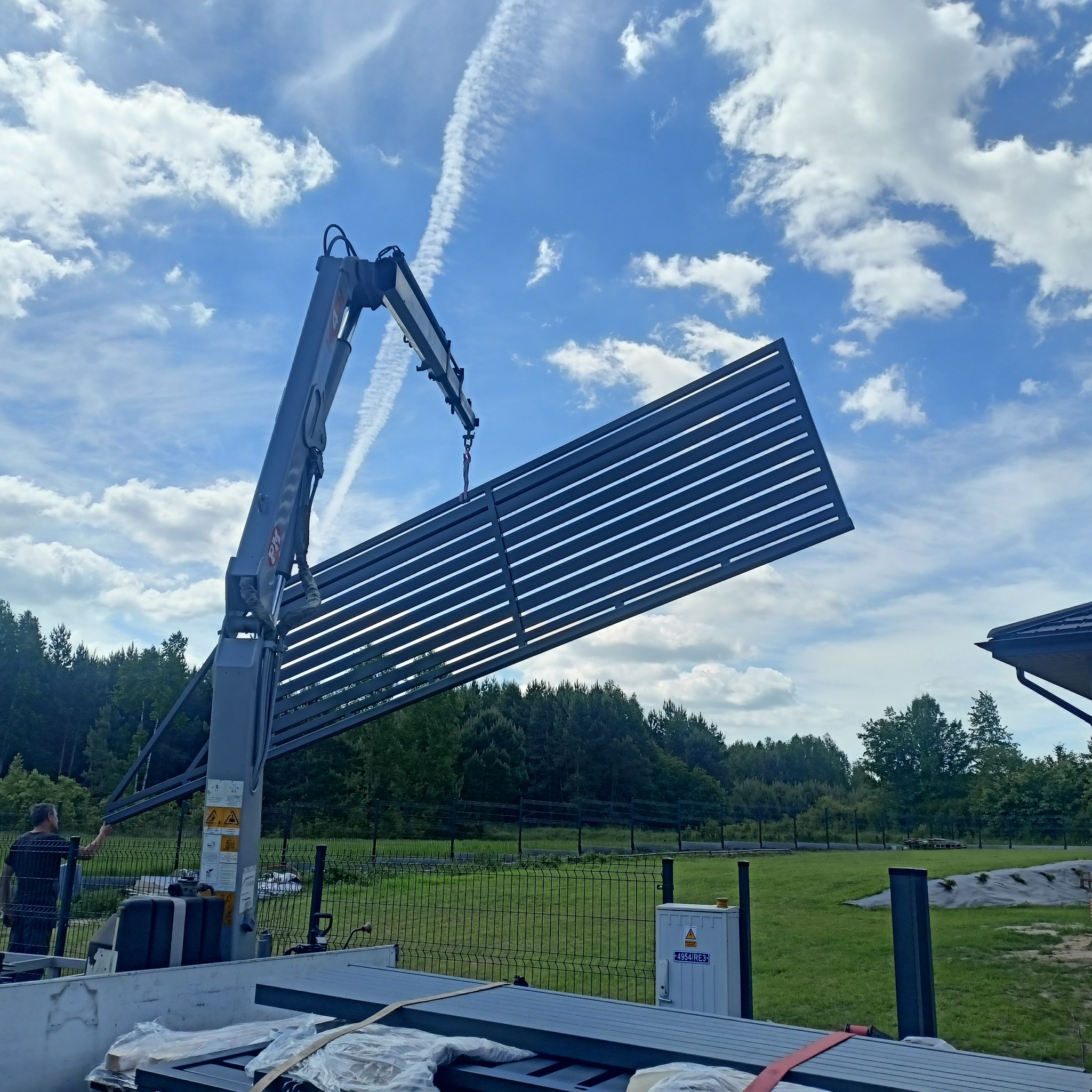 Montaż nowoczesnego ogrodzenia panelowego z użyciem dźwigu na tle błękitnego nieba. Widoczny pracownik nadzorujący proces i elementy ogrodzenia przygotowane do montażu.