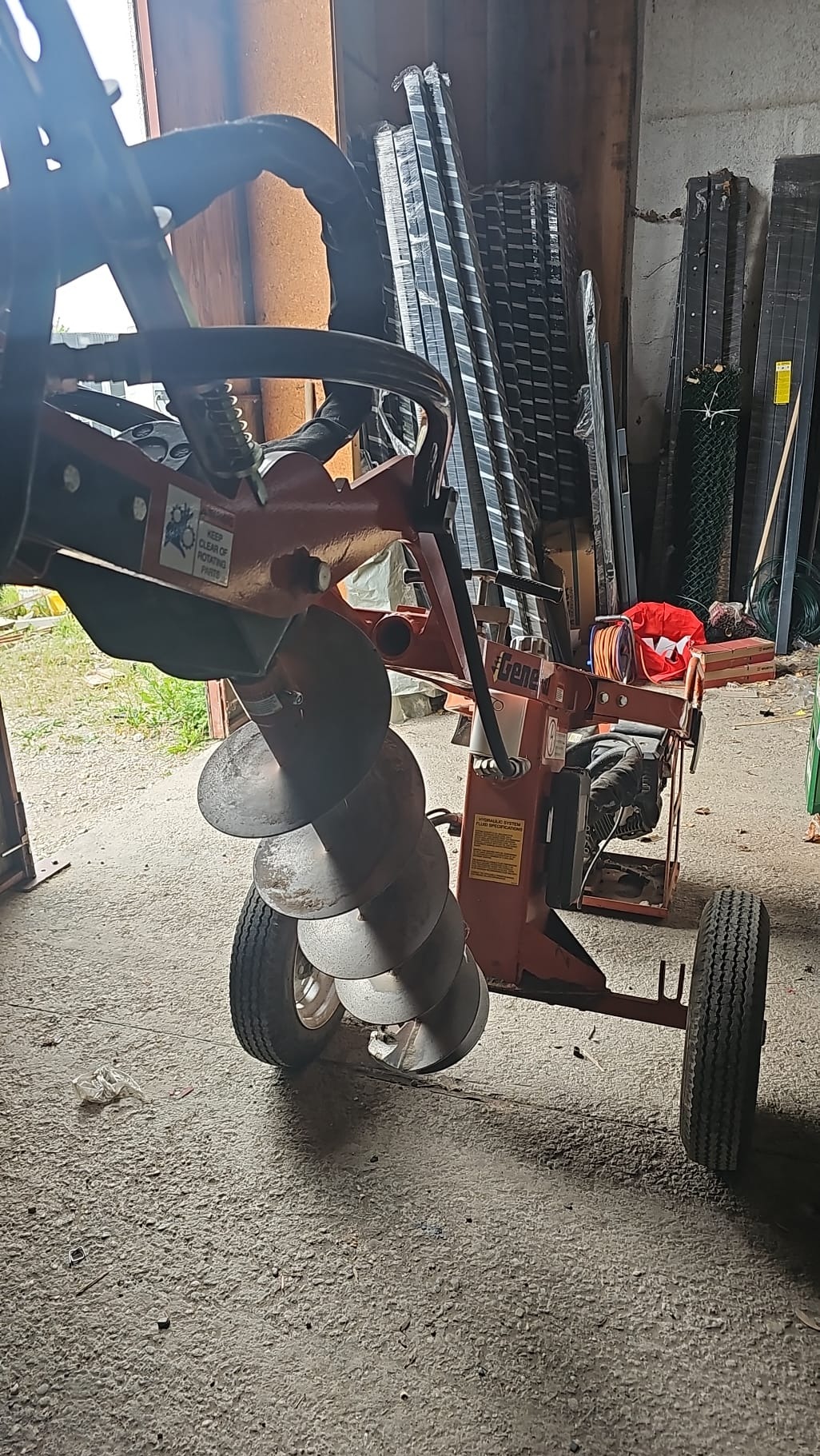 Wiertnica glebowa na kółkach, z widocznym świdrem, w tle ogrodzenia panelowe i siatka ogrodzeniowa. Sprzęt do przygotowania podłoża pod montaż.