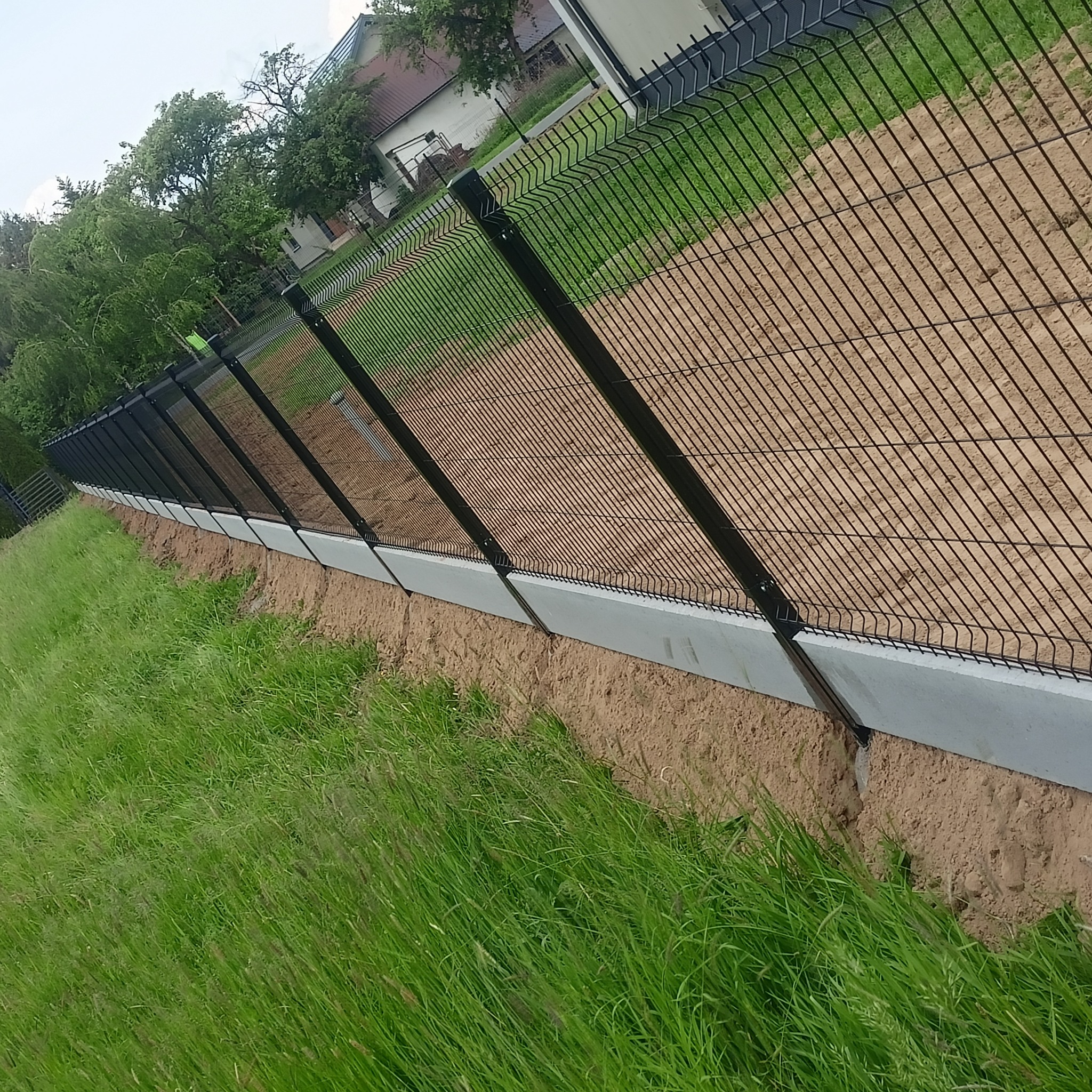 Czarna siatka ogrodzeniowa panelowa, zamontowana na betonowej podmurówce, oddziela trawnik od terenu z piaskiem, w tle dom.