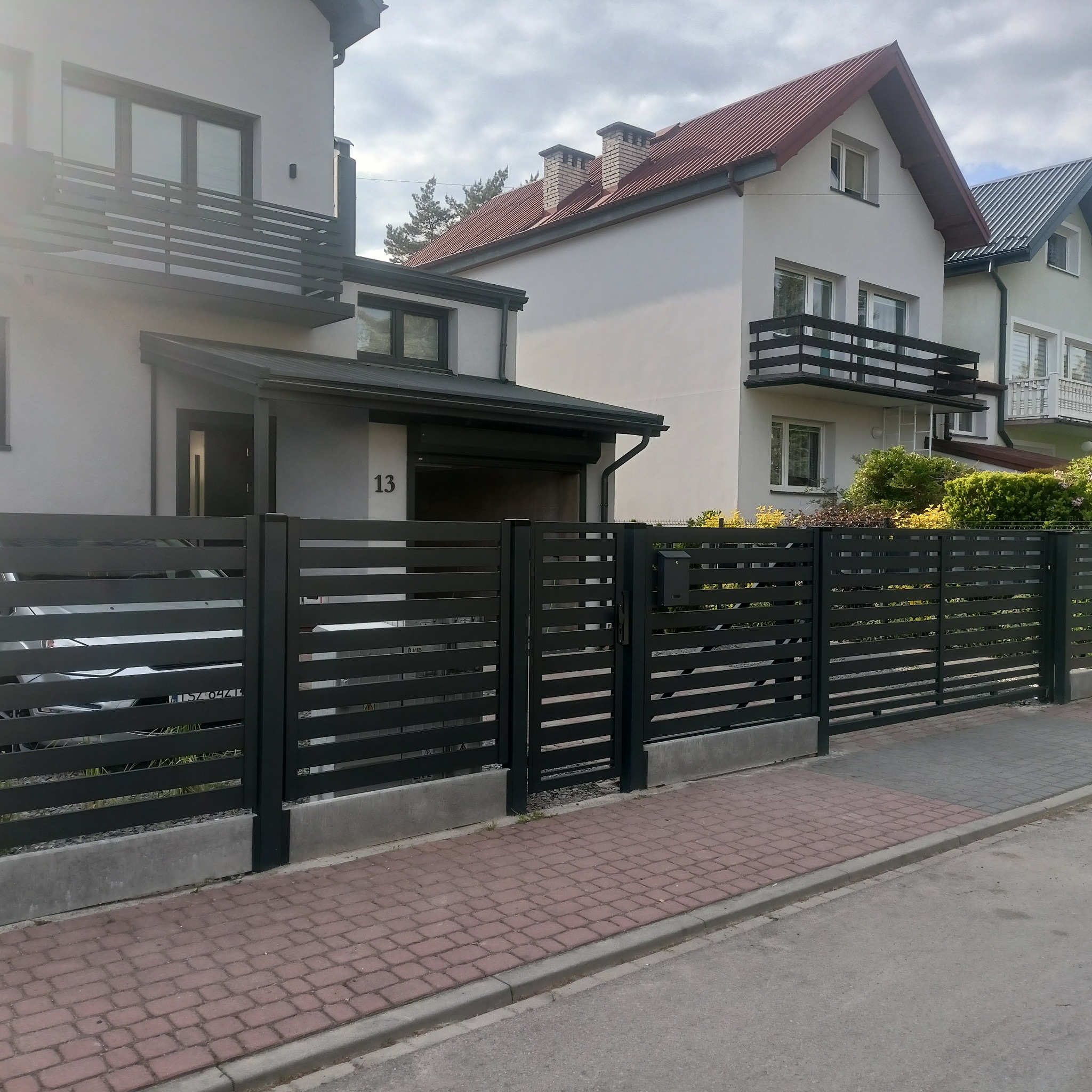 Nowoczesne ogrodzenie posesji z poziomych, grafitowych paneli, na tle jasnych budynków mieszkalnych. Widoczna brama i furtka.