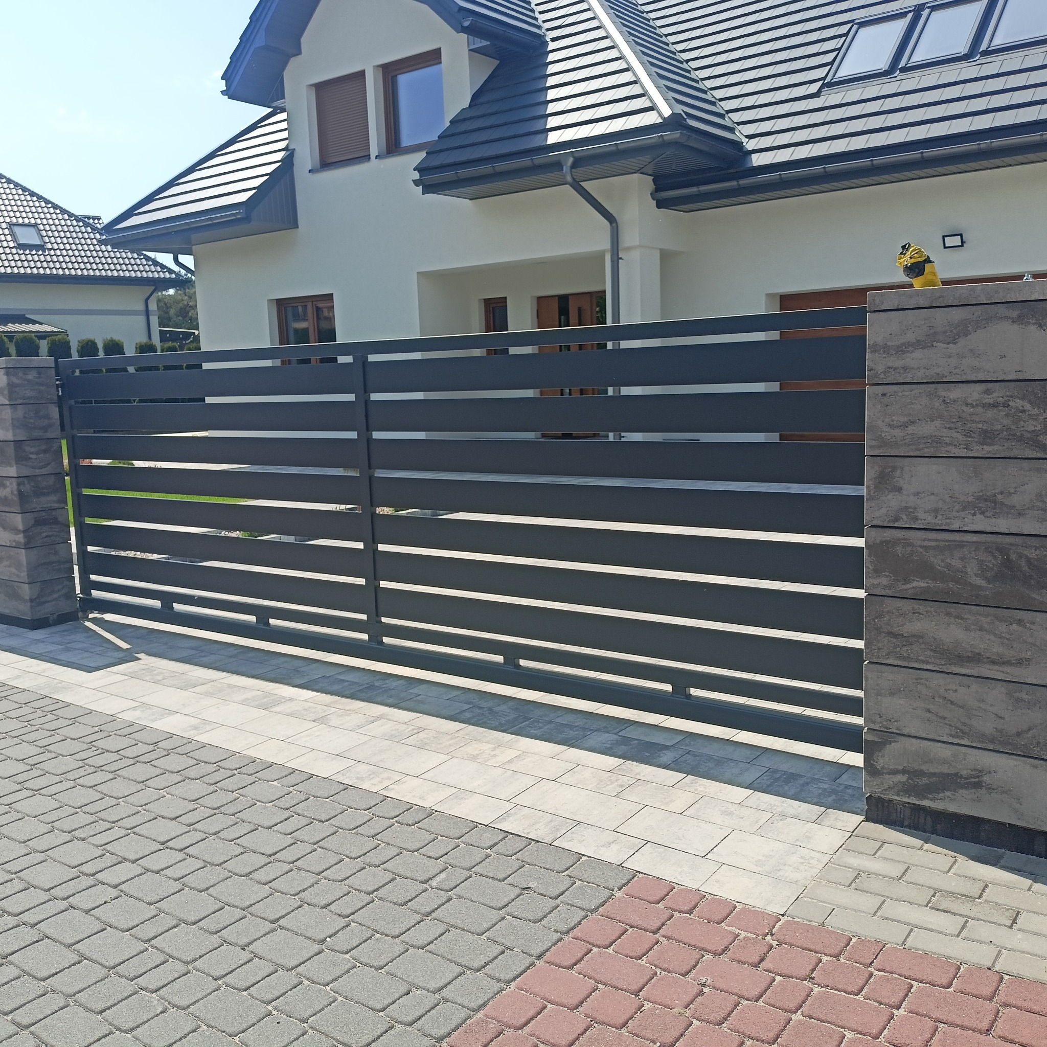 Nowoczesna, pozioma brama wjazdowa w kolorze antracytu, z elementami z szarego betonu architektonicznego, na tle domu z ciemną dachówką i kostki brukowej.