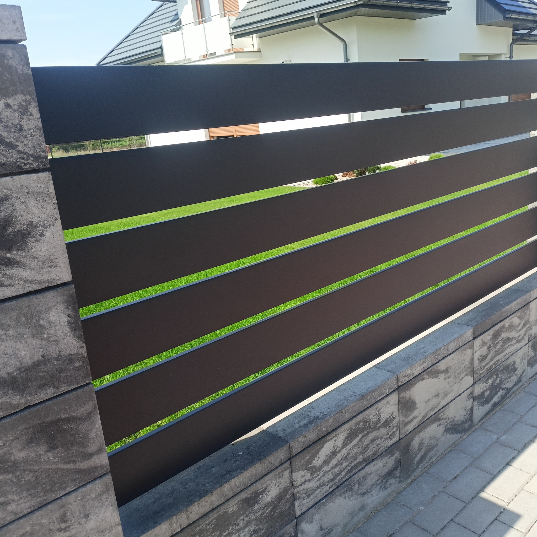 Nowoczesne ogrodzenie z poziomych, grafitowych paneli na betonowej podmurówce. W tle widoczny dom z białymi ścianami i szarym dachem.