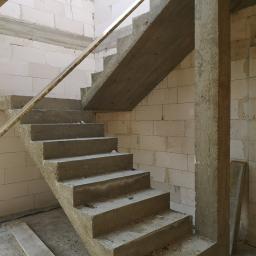 ADM - Surowe betonowe schody w trakcie budowy, wsparte na betonowym filarze, widoczne białe ściany z bloczków, brak wykończenia.