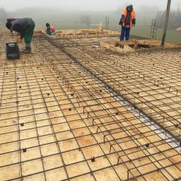 ADM - Przygotowanie szalunku i zbrojenia płyty fundamentowej na budowie w pochmurny dzień; widoczni pracownicy budowlani.