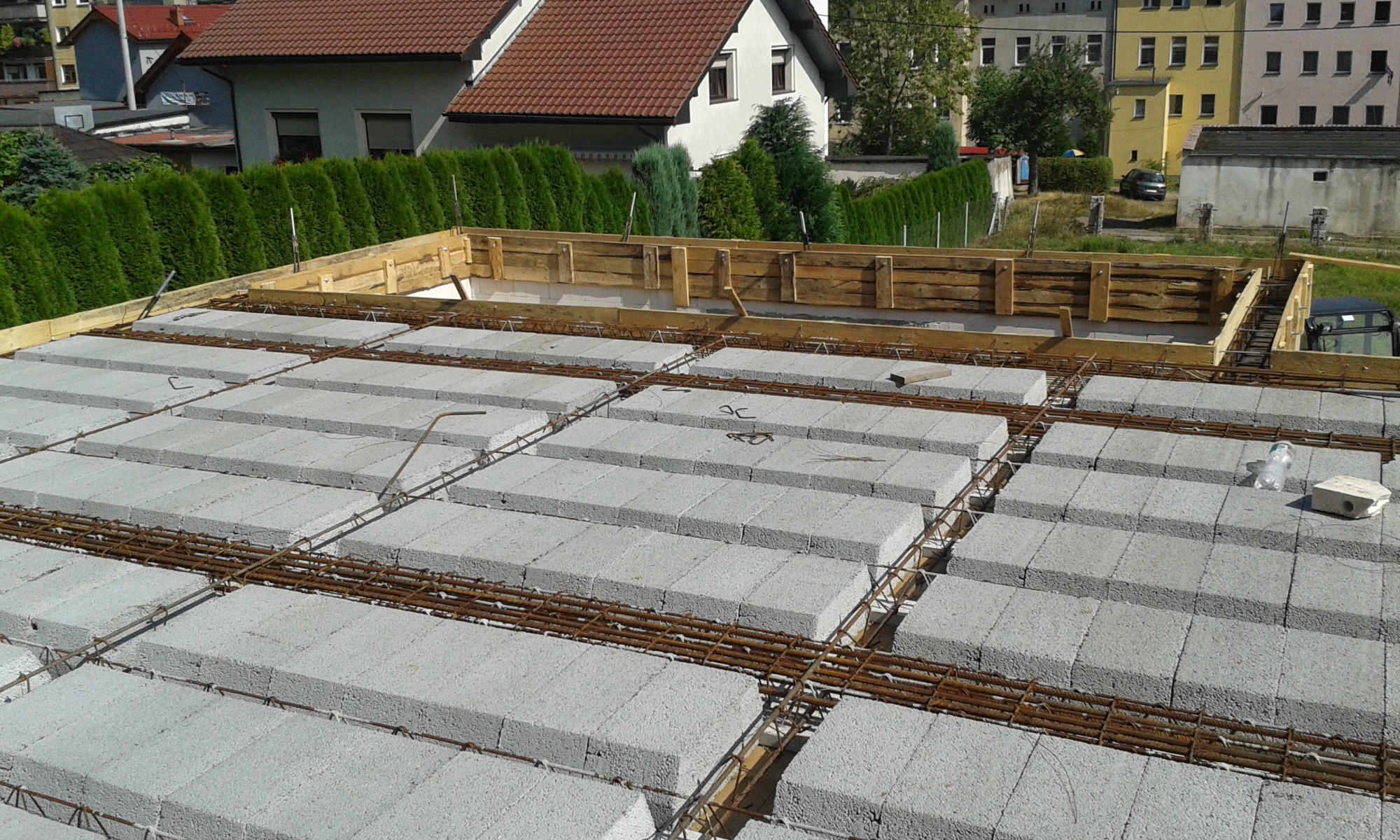 Wiązanie zbrojenia na stropie z pustaków szalunkowych, widoczne drewniane szalunki obwodowe i zabudowa jednorodzinna w tle.