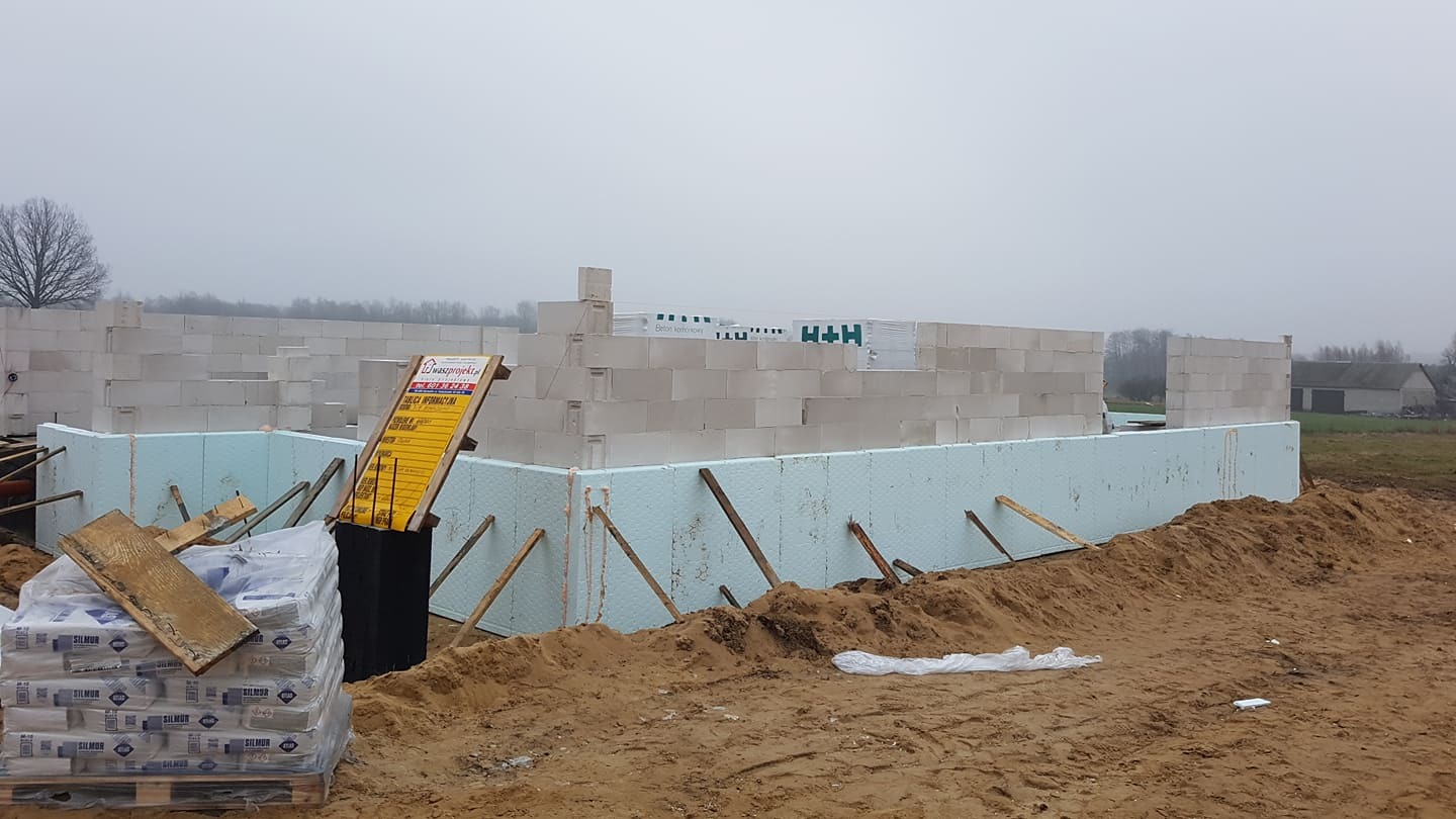 Budowa domu jednorodzinnego w trakcie wznoszenia ścian z bloczków betonowych, widoczne ocieplenie fundamentów i stos worków z zaprawą, tablica informacyjna budowy po lewej stronie.