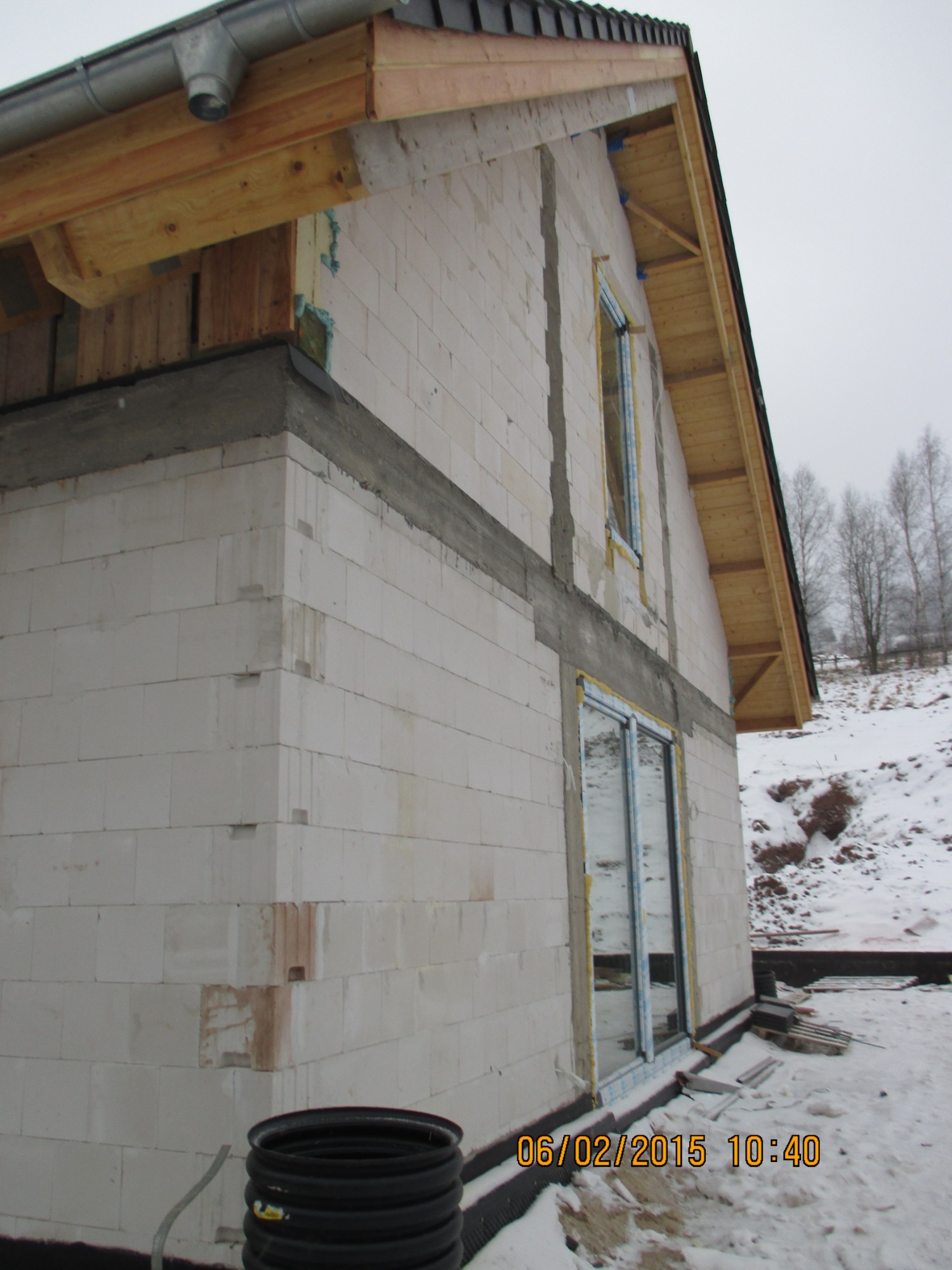 Surowy stan budynku w trakcie budowy: ściany z białych bloczków, wstawione okna, widoczna konstrukcja dachu, zimowy krajobraz w tle.