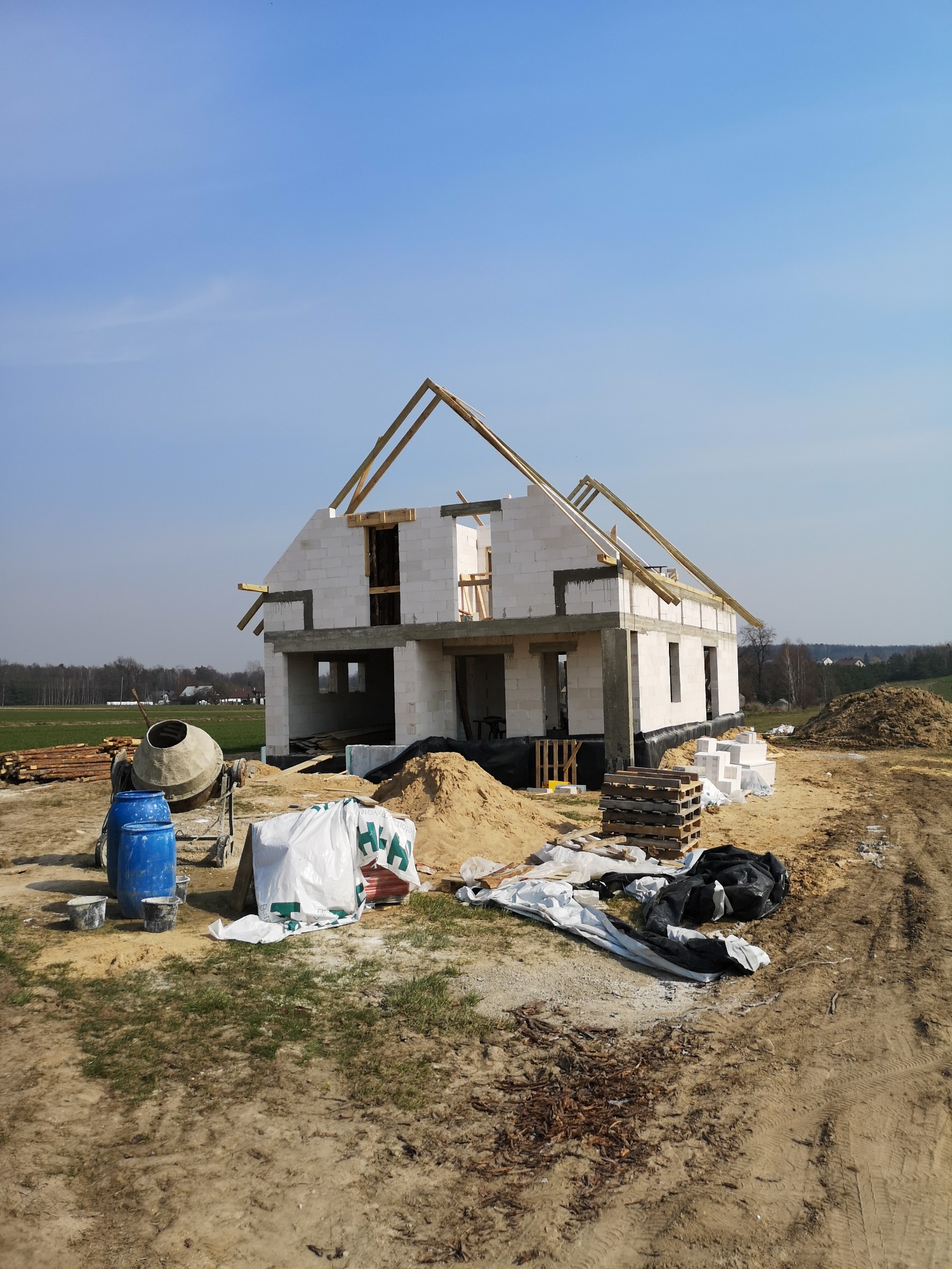 Budowa domu jednorodzinnego w stanie surowym otwartym, widoczne ściany z bloczków, więźba dachowa, mieszarka do betonu i materiały budowlane na placu budowy w słoneczny dzień.
