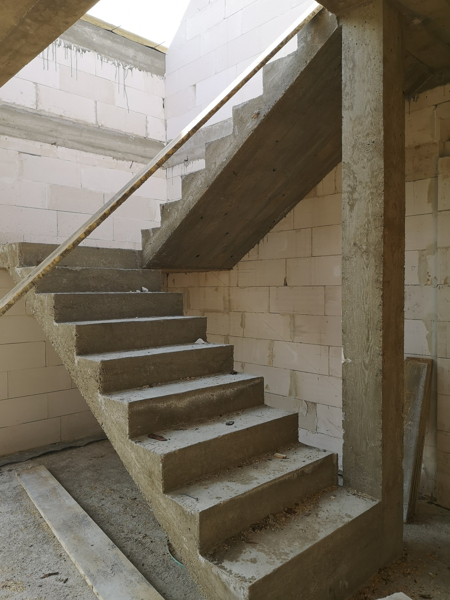 Surowe betonowe schody w trakcie budowy, wsparte na betonowym filarze, widoczne białe ściany z bloczków, brak wykończenia.