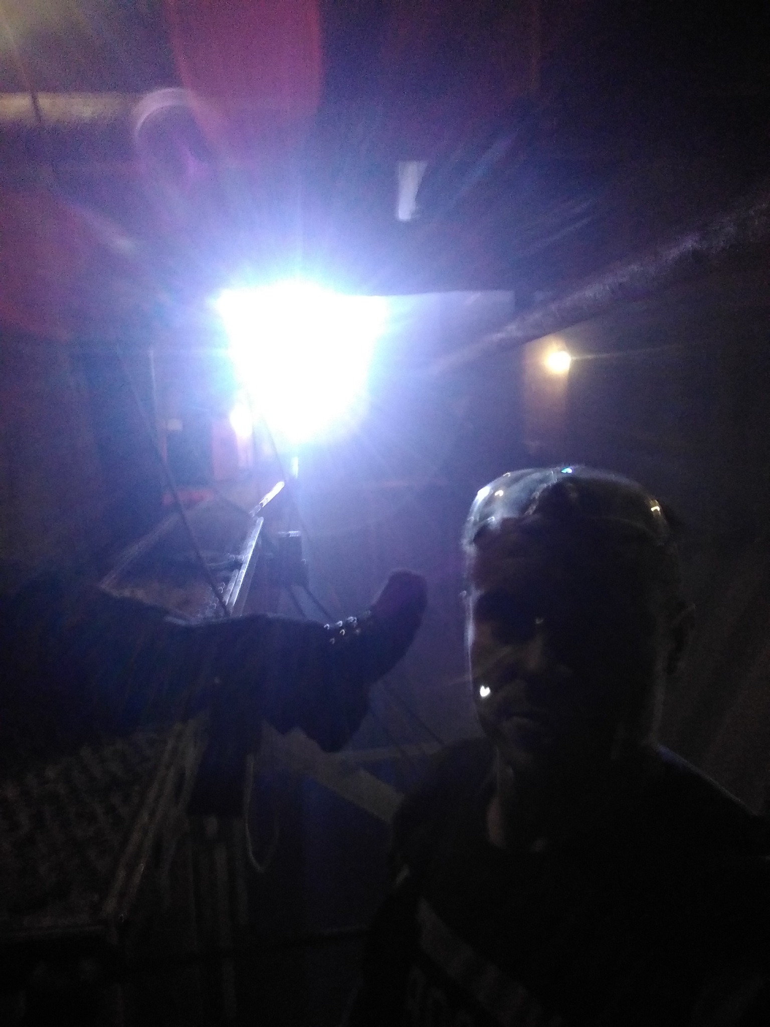 Słabo oświetlone wnętrze z fragmentem drabiny, pracownikiem w ciemnej odzieży i silnym źródłem światła w tle.