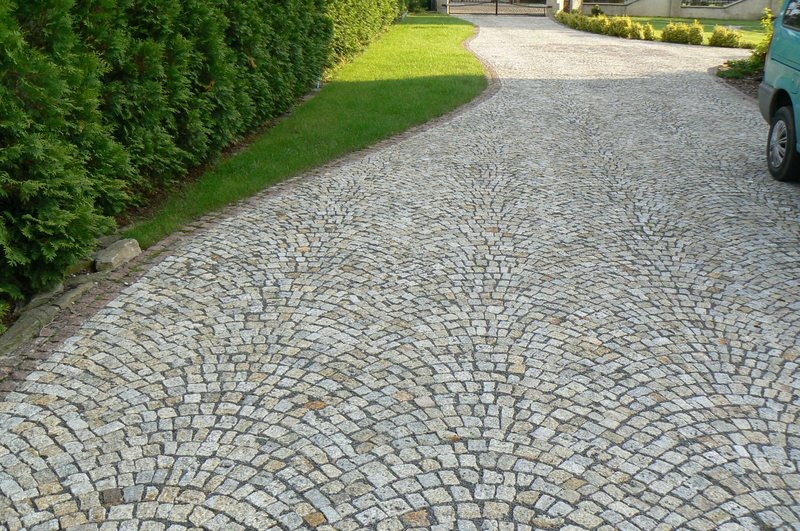 Podjazd z kostki granitowej ułożonej w wachlarz, obrzeża z kostki, po lewej żywopłot z tui, w tle brama wjazdowa.