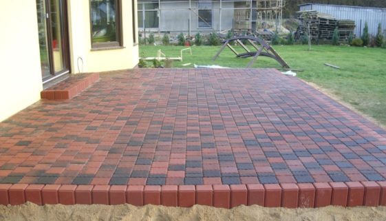 Ułożony taras z kostki brukowej w odcieniach czerwieni i grafitu, otoczony obrzeżem z czerwonej kostki. Widoczny fragment budynku z oknem i trawnik w tle.