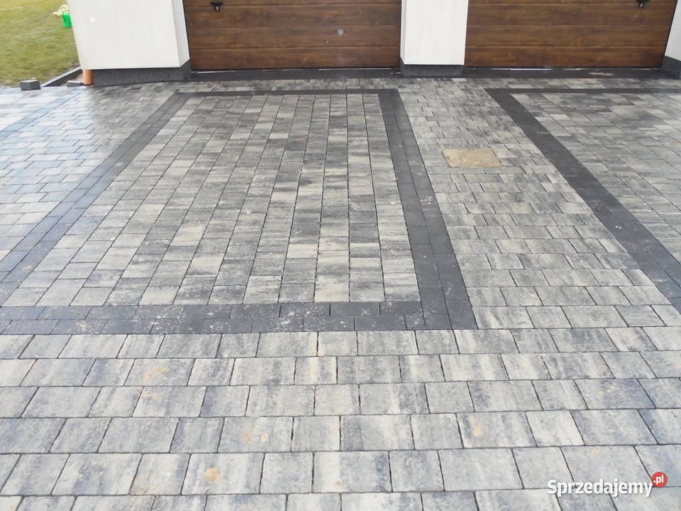 Wykończony podjazd z kostki brukowej w odcieniach szarości, obramowany ciemniejszą kostką, prowadzący do garażu z drewnianymi drzwiami.