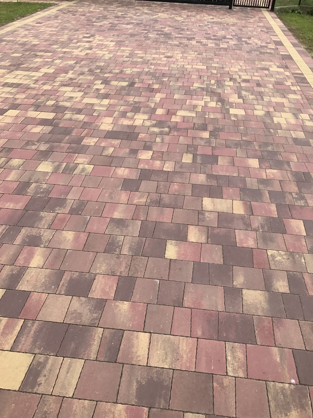 Ułożona kostka brukowa w odcieniach brązu, beżu i czerwieni tworząca wzór na podjeździe, widoczna brama w tle.