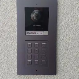 Szary, modułowy wideodomofon HikVision z panelem numerycznym i przyciskiem dzwonka na teksturowanej, jasnej ścianie zewnętrznej.