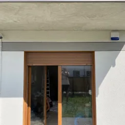 Zewnętrzny montaż zabezpieczeń: kamera i sygnalizator nad oknem z roletami, widoczne okablowanie i czujnik ruchu. W odbiciu szyby widoczna osoba robiąca zdjęcie oraz wnętrze z drabiną.