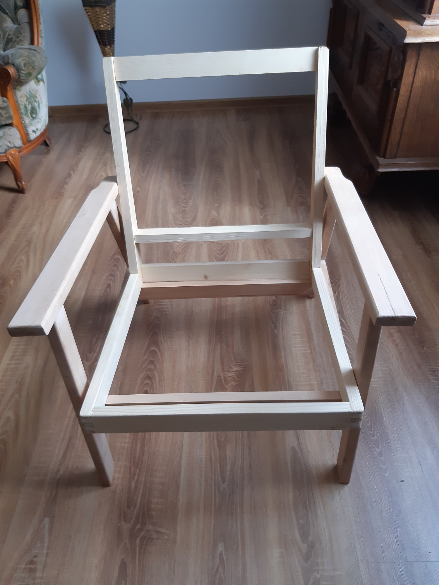 Surowy, drewniany szkielet fotela z podłokietnikami, widoczny na tle drewnianej podłogi, w trakcie procesu tworzenia.