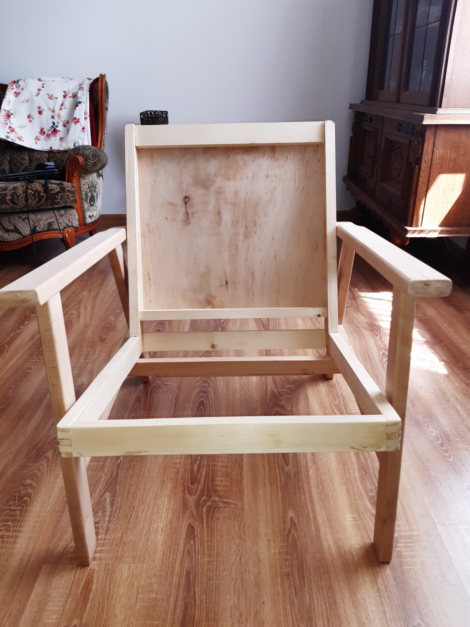 Surowy, drewniany szkielet fotela z widocznymi połączeniami stolarskimi, na tle parkietu i antycznych mebli.