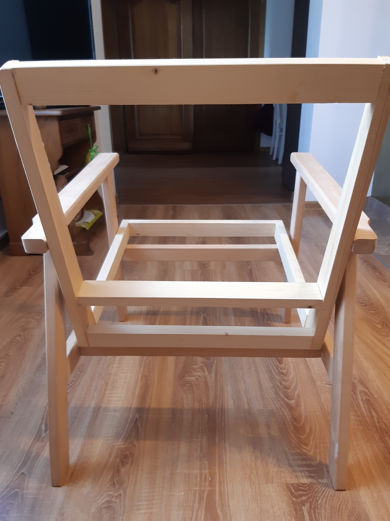 Surowa drewniana rama krzesła z podłokietnikami, widok z tyłu, ukazująca konstrukcję i połączenia stolarskie, na tle parkietu.