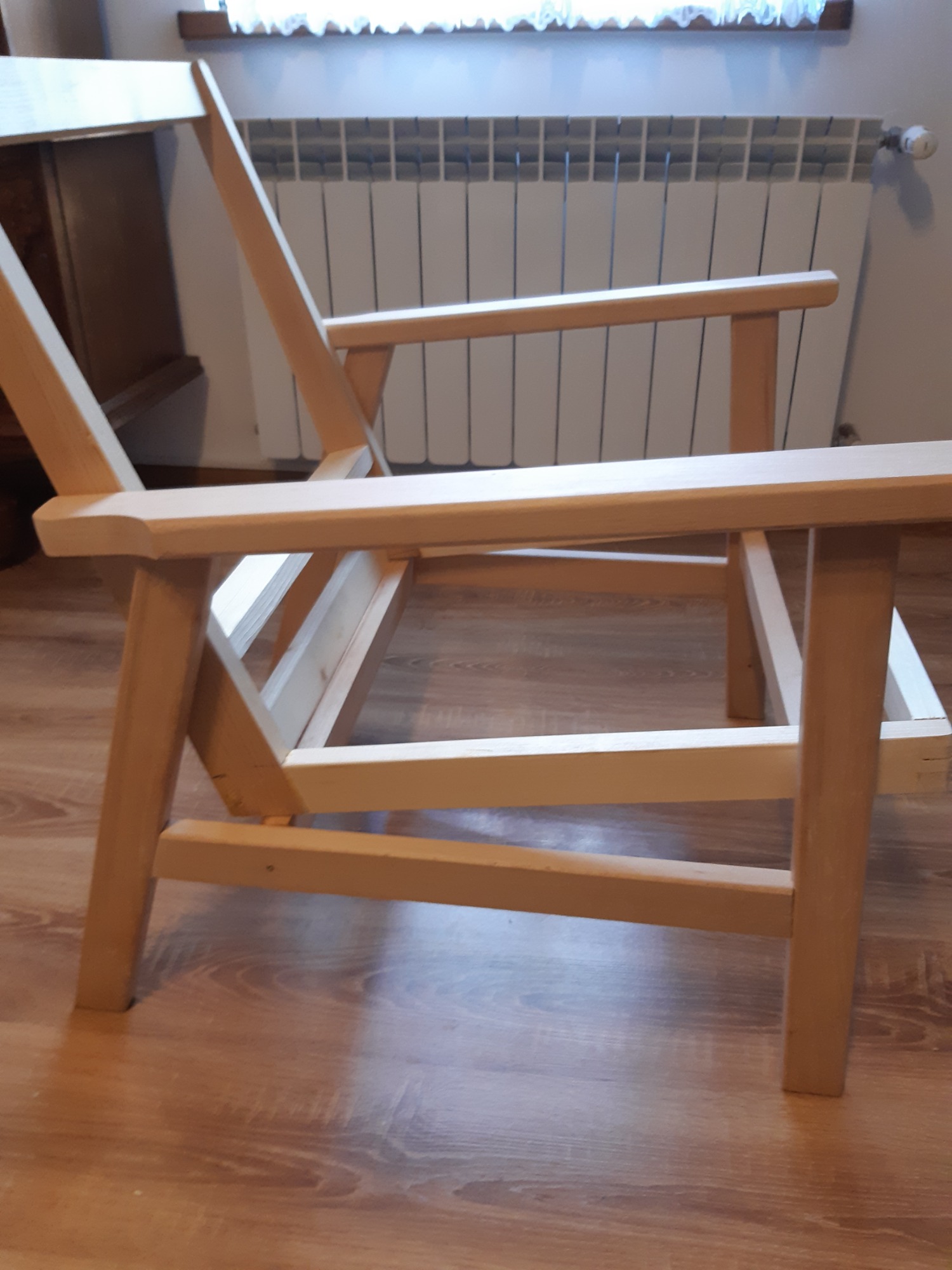 Surowe drewniane krzesło z podłokietnikami, niedokończone, na tle grzejnika i okna z firanką.