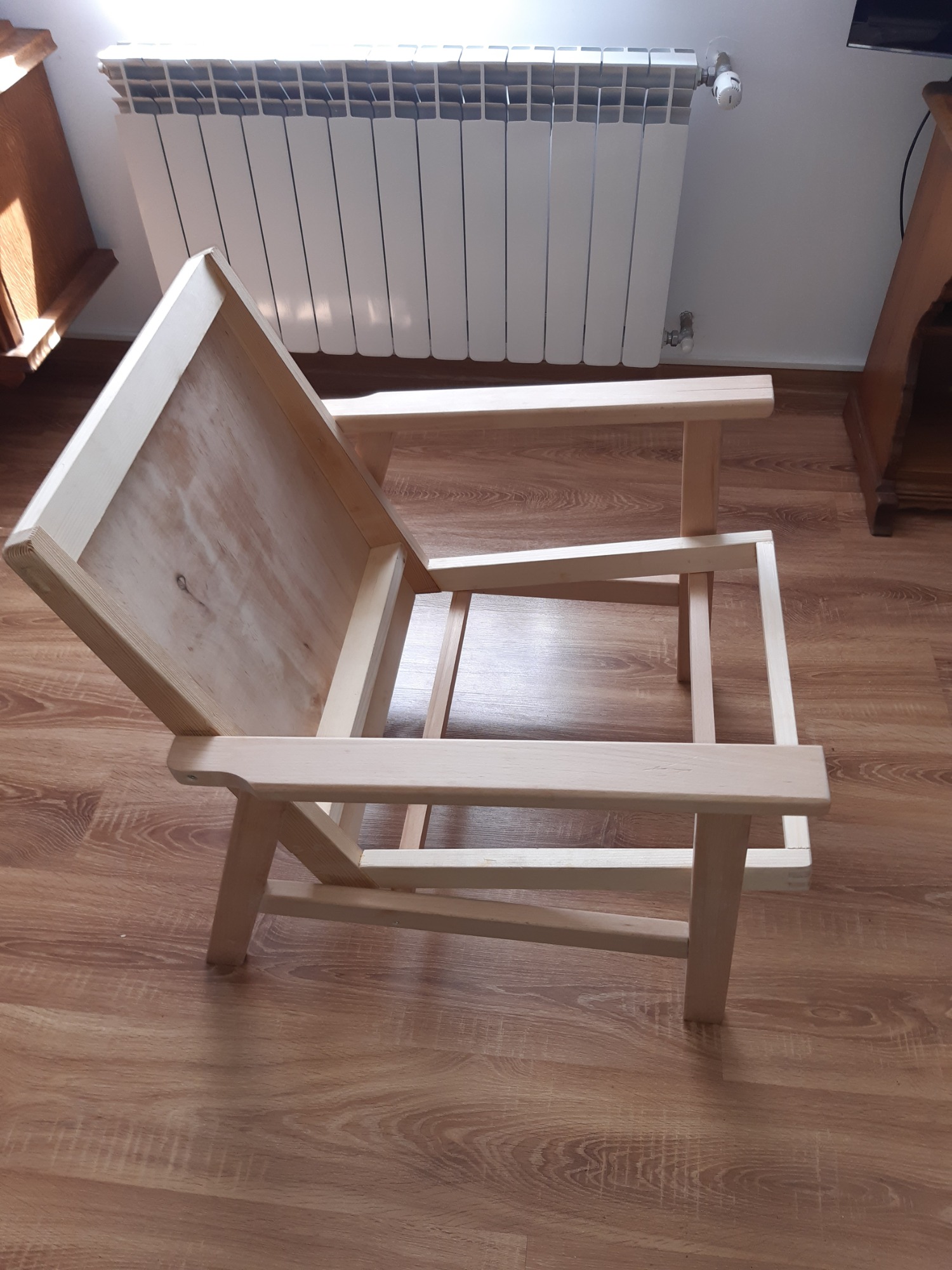 Surowy, drewniany szkielet krzesła z podłokietnikami, ustawiony na panelach podłogowych, z grzejnikiem i fragmentami innych mebli w tle.