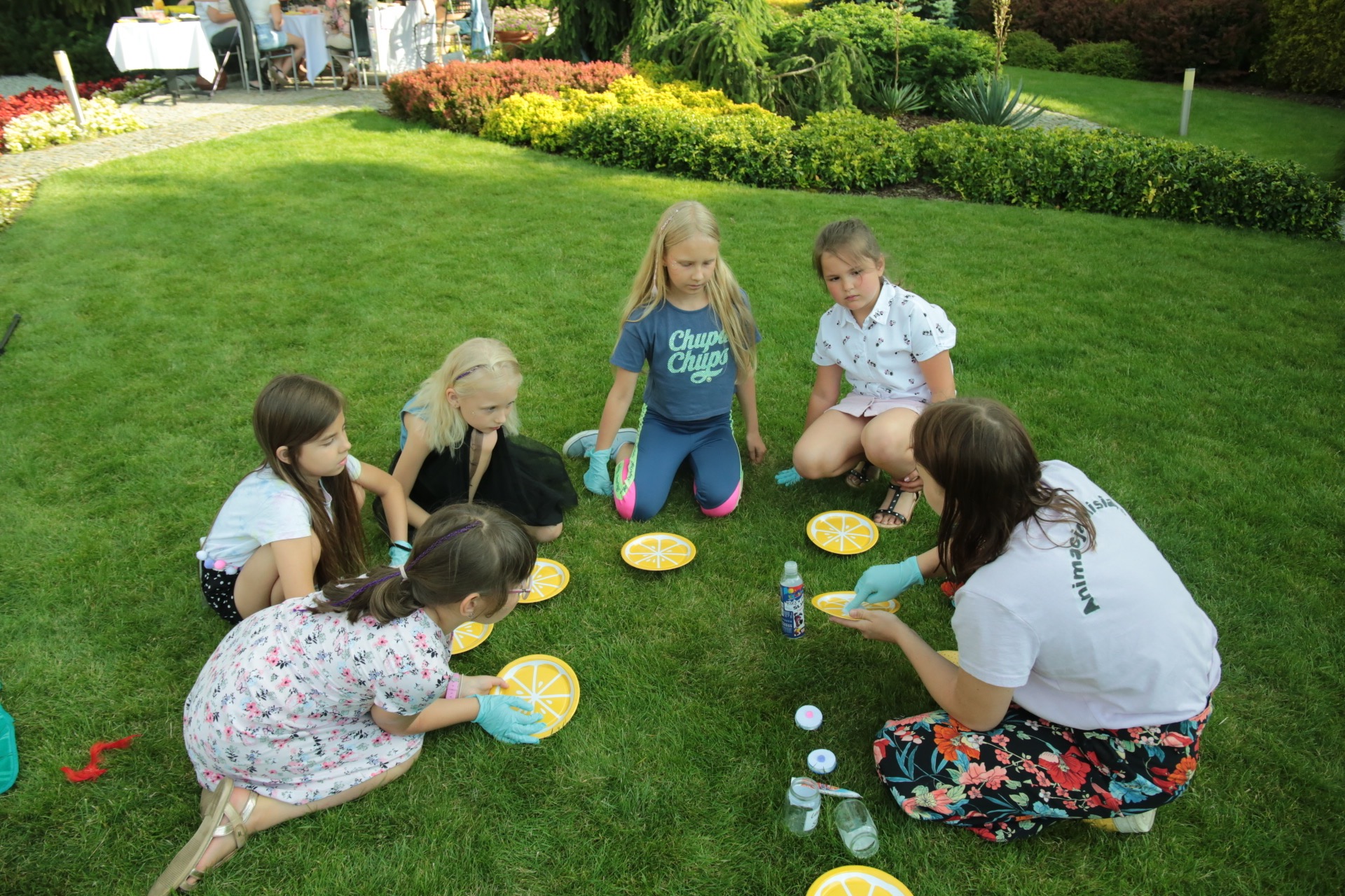 Grupa dzieci w ogrodzie, uczestnicząca w warsztatach plastycznych na trawie, malując farbami na papierowych talerzach w kształcie pomarańczy pod okiem animatorki w koszulce z napisem 'Animacja'.