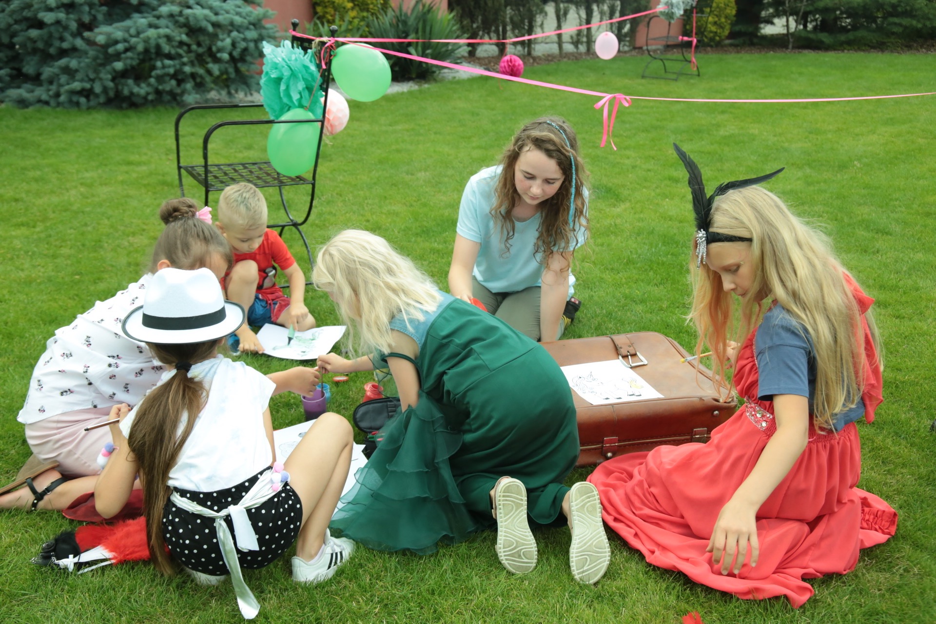 Grupa dzieci siedzi na trawie, kolorując obrazki; dekoracje ogrodowe, balony i rekwizyty rozłożone wokół nich, tworząc radosną atmosferę.