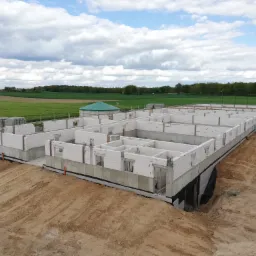 Wznoszenie ścian parteru domu z białych bloczków, widoczne zbrojenia, izolacja fundamentów, w tle zielone pola i błękitne niebo z chmurami.