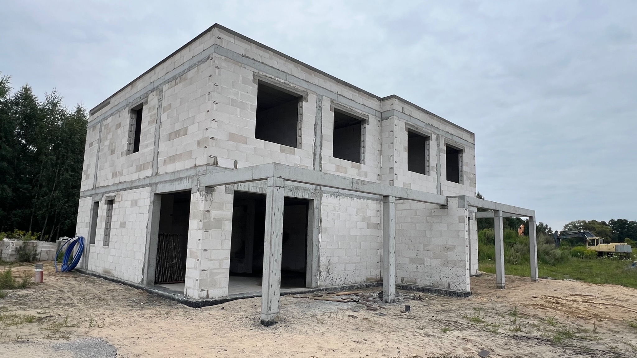 Surowy stan dwukondygnacyjnego domu z betonu komórkowego, widoczne otwory okienne i drzwiowe, betonowe nadproża, teren budowy z piaskiem i sprzętem w tle.