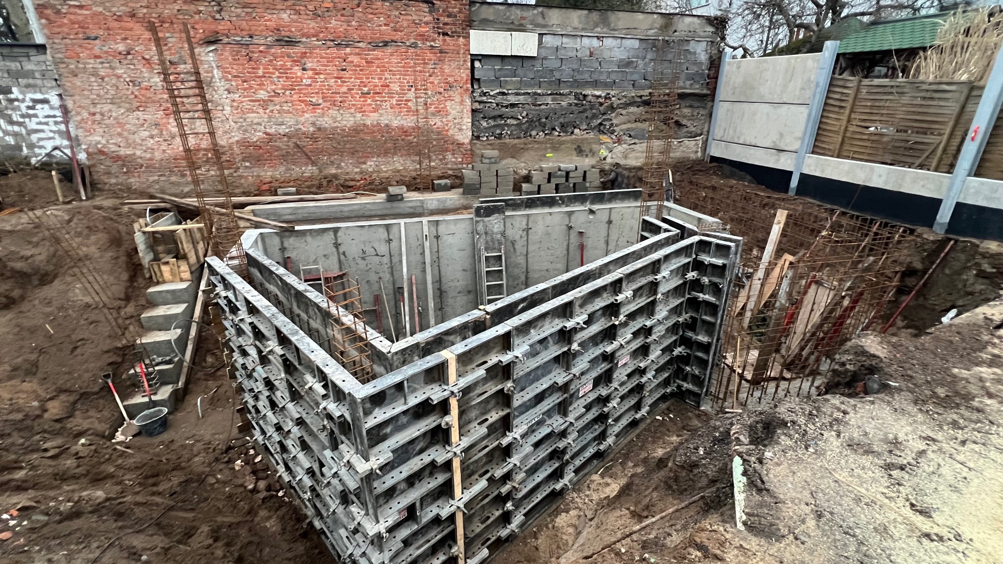 Szare szalunki fundamentowe w wykopie, widok z góry. W tle ceglany budynek i betonowe ściany. Elementy zbrojenia i drabiny w wykopie.