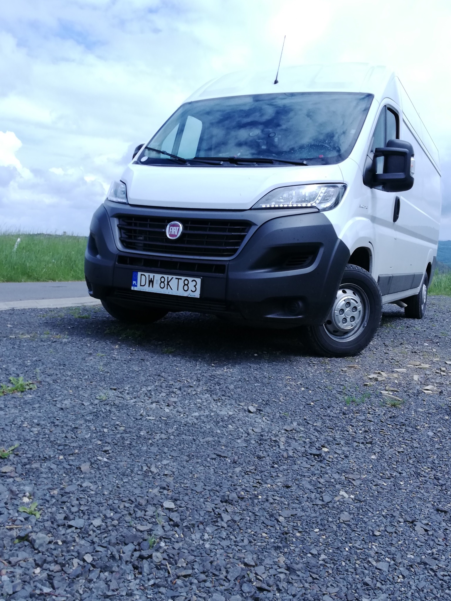 Biały bus marki Fiat Ducato z polską tablicą rejestracyjną DW 8KT83 zaparkowany na żwirowym podłożu, z zielonymi wzgórzami w tle i pochmurnym niebem.