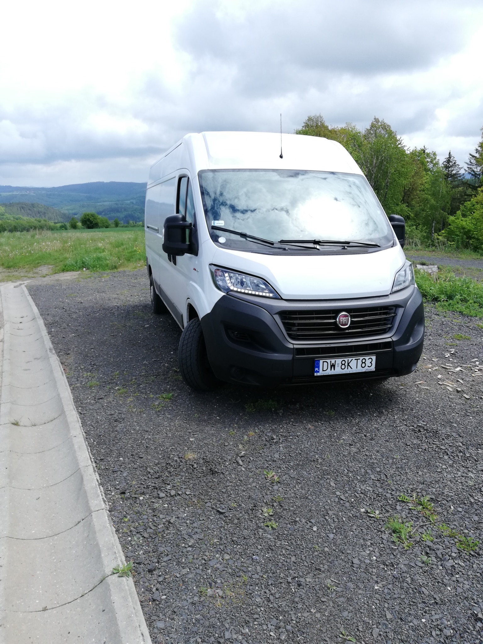Biały van Fiat Ducato zaparkowany na żwirowym placu z widokiem na zalesione wzgórza w tle.