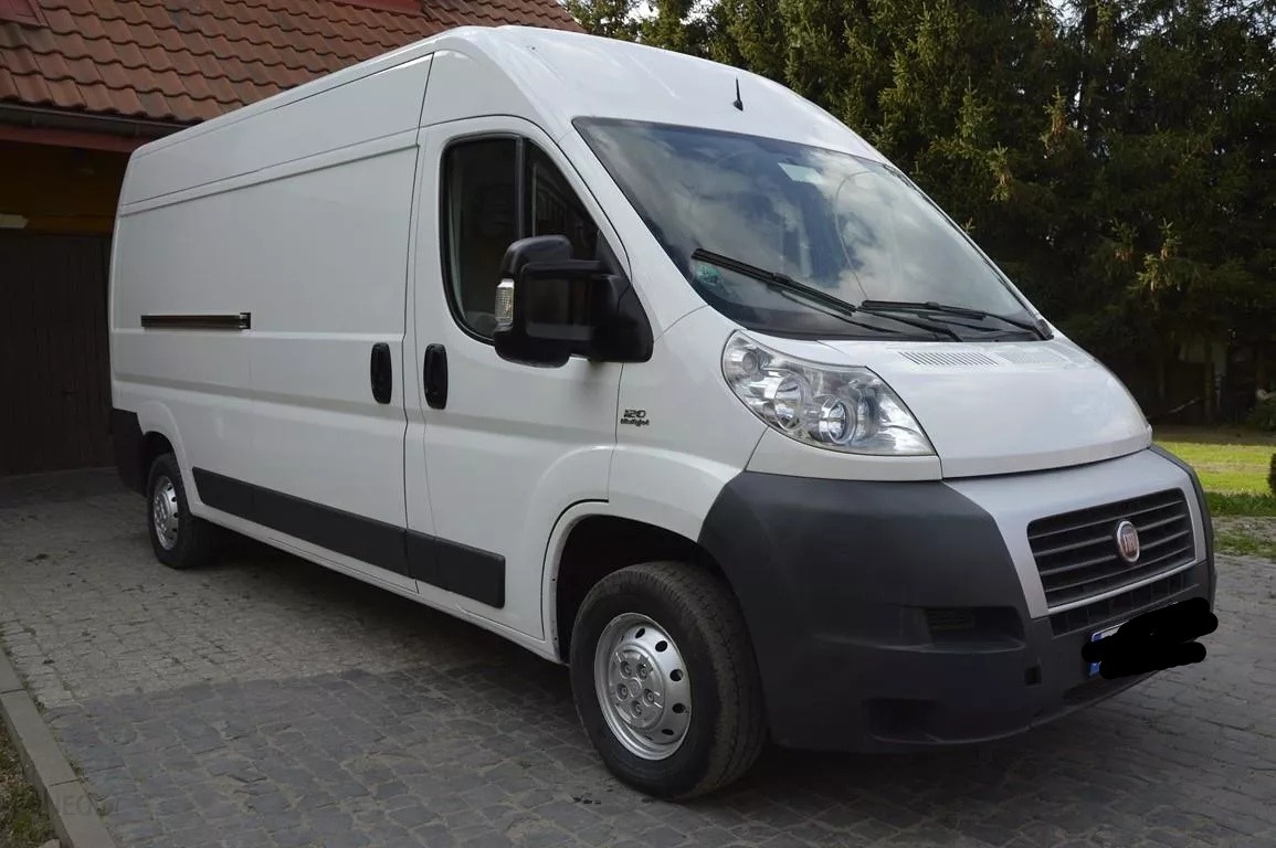 Biały, dostawczy Fiat Ducato 120 Multijet zaparkowany na brukowanej nawierzchni przed budynkiem z czerwoną dachówką i zielonymi drzewami w tle.