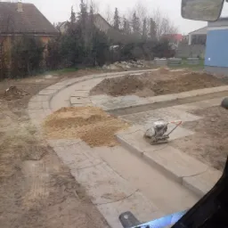 Przygotowany teren pod ścieżkę z ułożonymi obrzeżami z kostki, ubijarka spalinowa na betonowym fundamencie, hałdy ziemi w tle, widok z kabiny pojazdu na plac budowy.