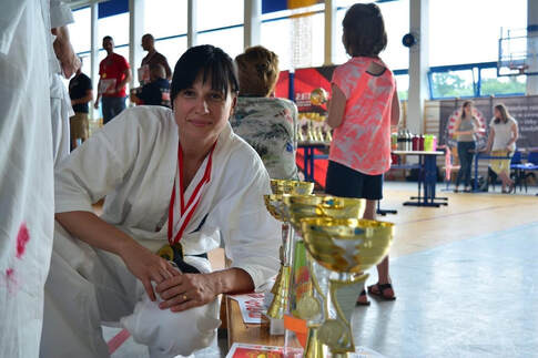 Uśmiechnięta kobieta w białym kimonie z medalem na szyi, siedząca przy stole z rzędem złotych pucharów w tle hali sportowej.