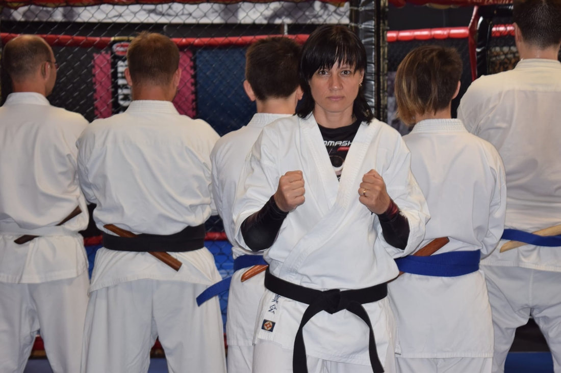 Instruktorka sportów walki w Gdańsku, w białym kimonie z czarnym pasem, prezentuje postawę bojową na tle grupy uczniów w kimonach z różnymi kolorami pasów.